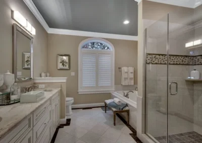 Elegant master bathroom with marble countertops, glass shower, soaking tub, and arched window with shutters.