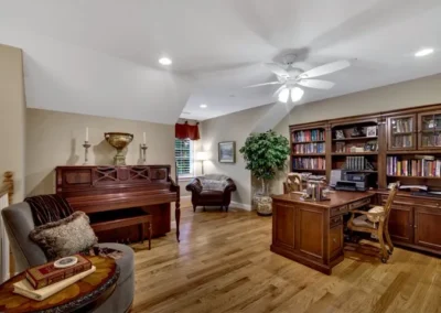 Cozy home office with wooden desk, bookshelves, piano, and armchair in well-lit room.