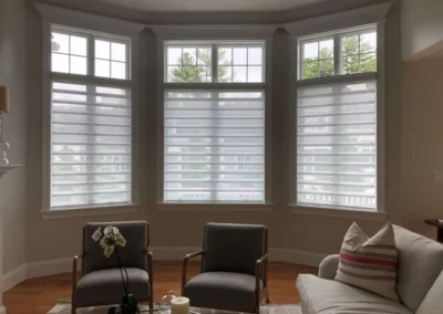 Bay window with sheer blinds, two gray chairs, orchid on table, light-filled living room.